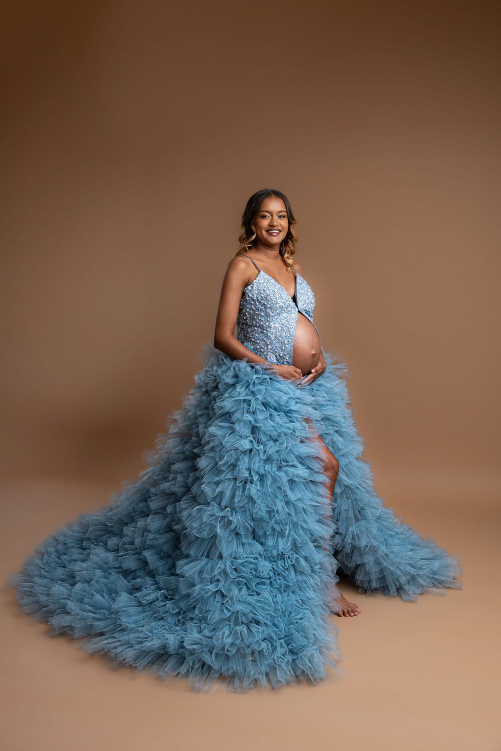 A maternity portrait in a flowing dusty blue tulle gown against a brown backdrop.