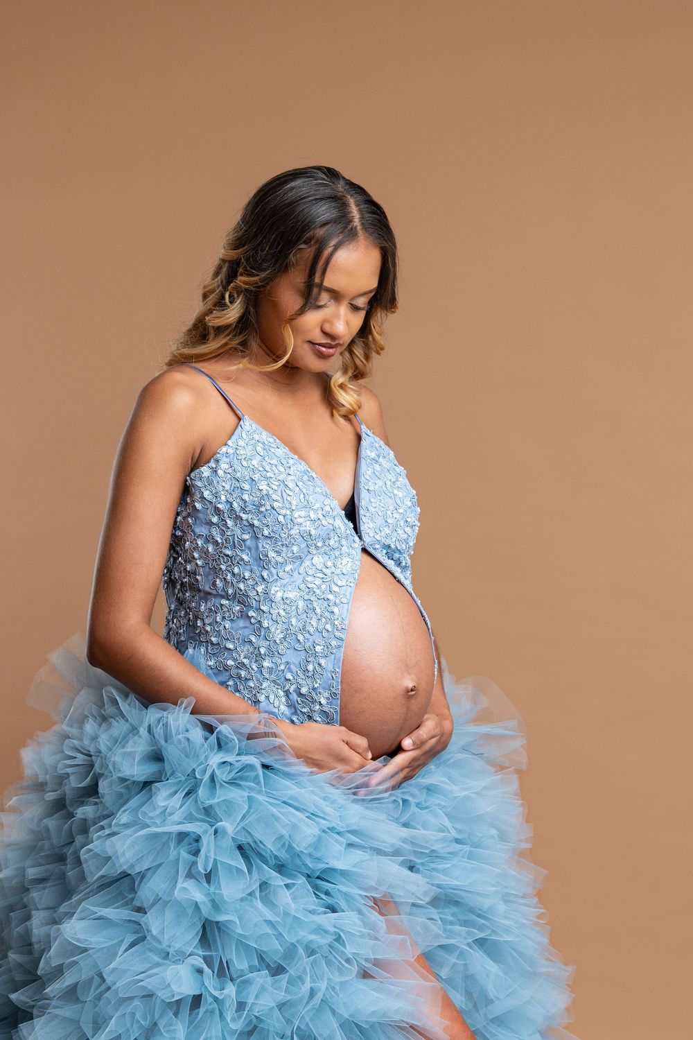 A pregnant woman in a light blue tulle gown poses gracefully against a neutral backdrop during a maternity photoshoot.