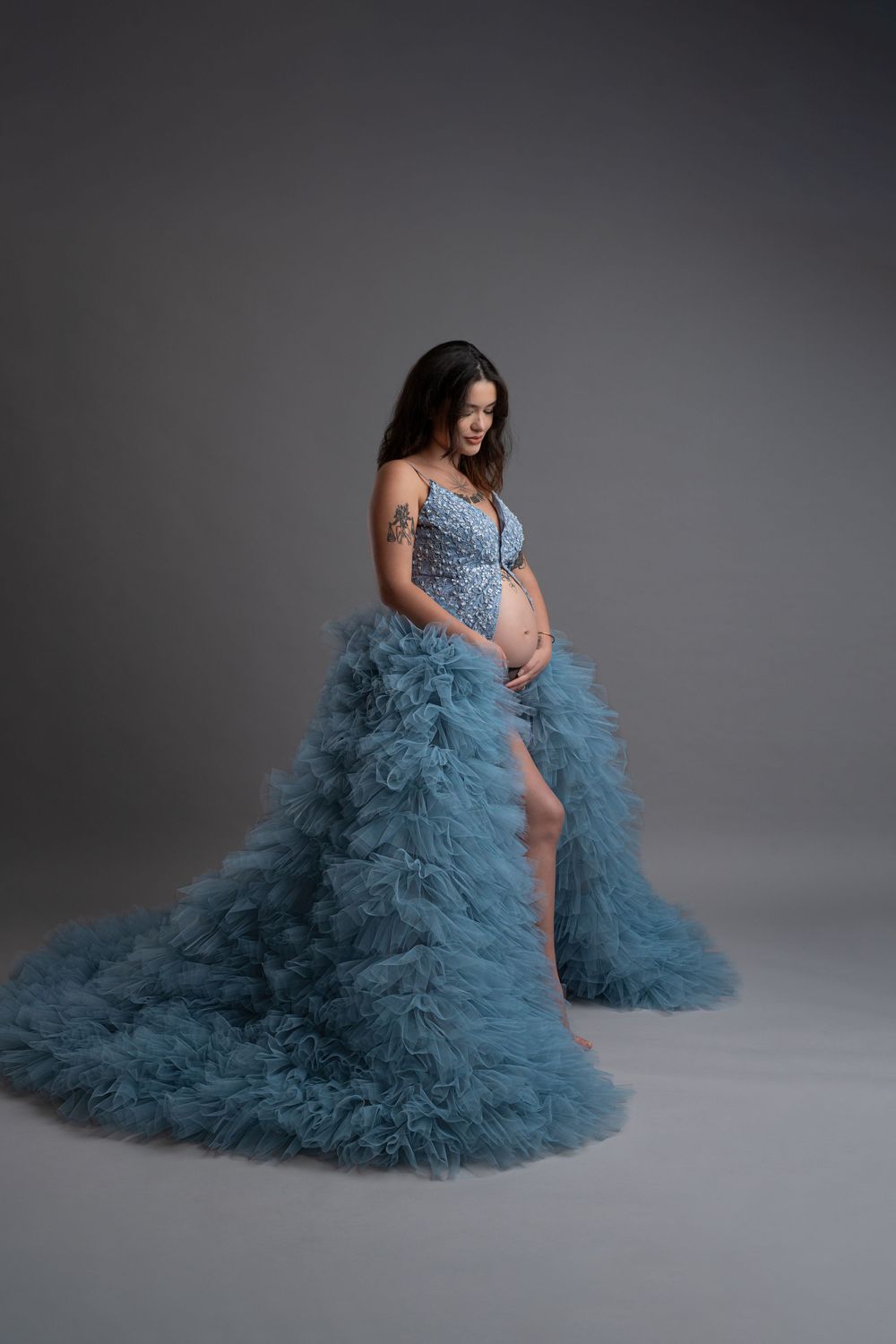 A maternity photo series showing a woman in a flowing dusty blue tulle gown against a gray studio backdrop.