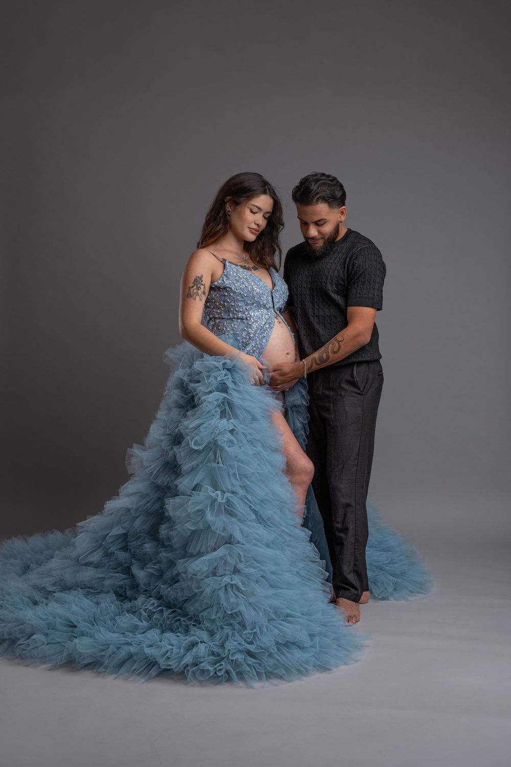 Couple in a studio setting with person wearing flowing dusty blue tulle gown while partner in black embraces them.