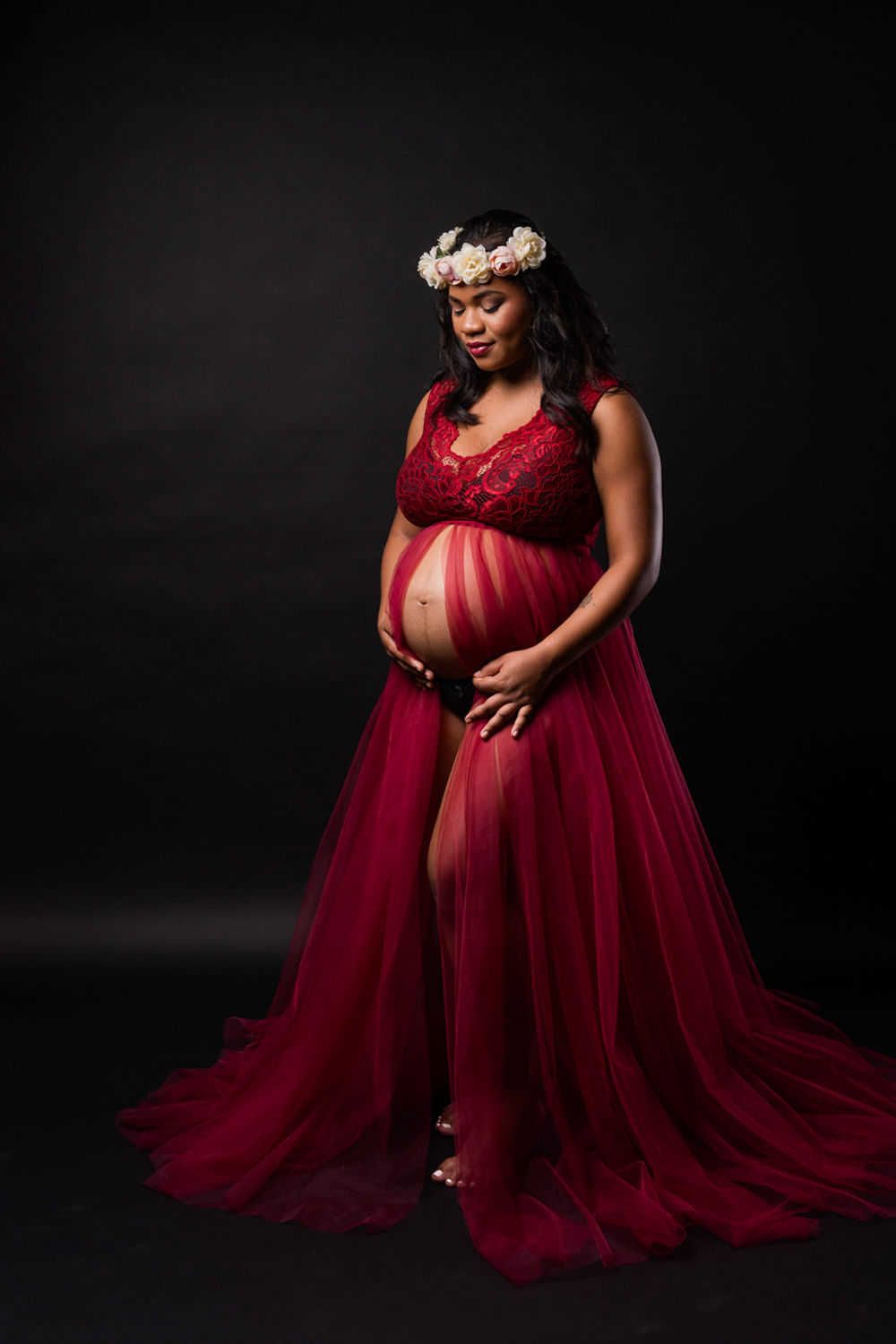 Portrait de maternité élégant mettant en vedette une robe fluide en tulle rouge bordeaux et une couronne florale sur un fond sombre.