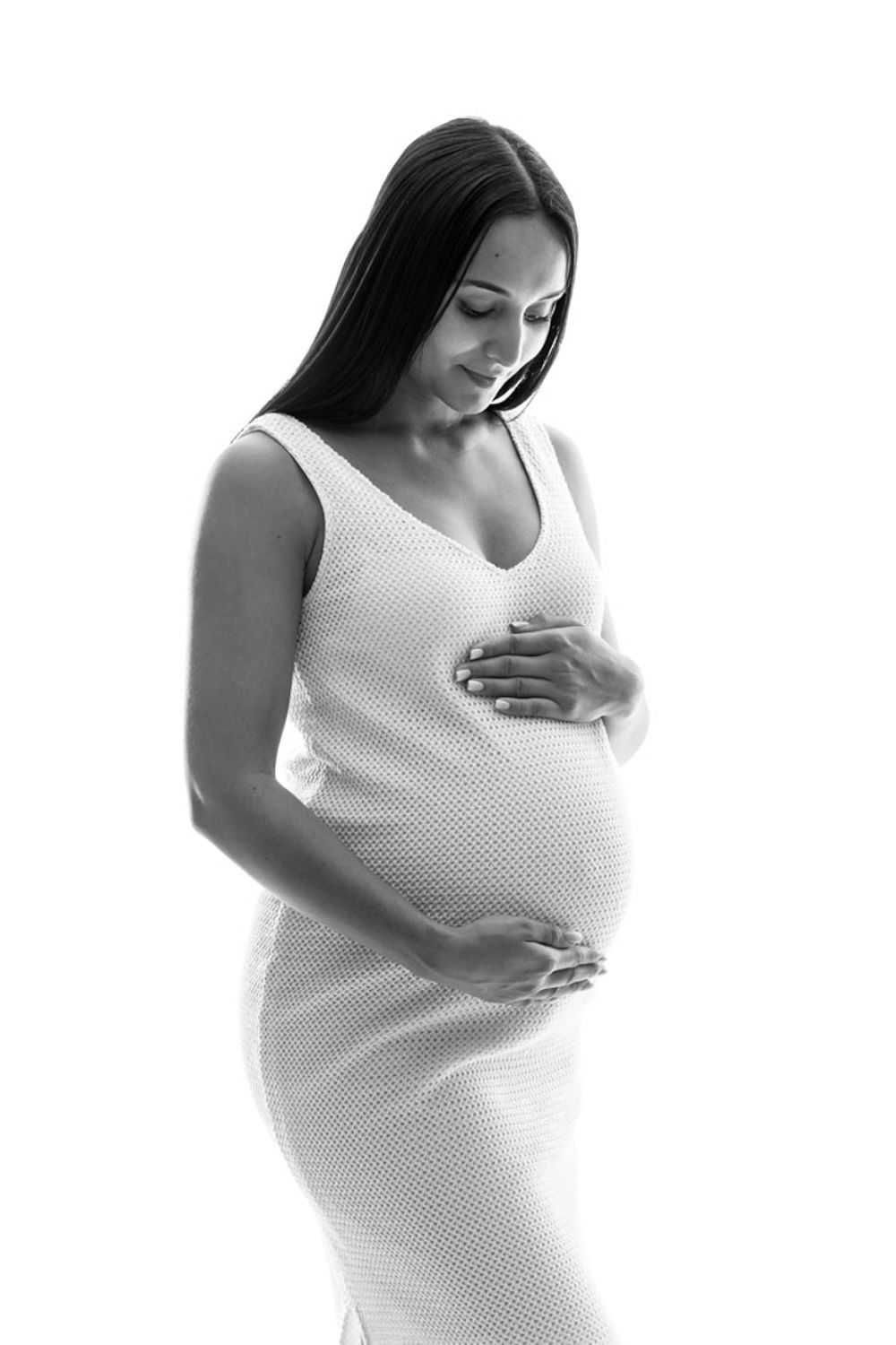 Portrait de maternité en noir et blanc d'une femme vêtue d'une robe blanche ajustée berçant doucement son ventre de femme enceinte.
