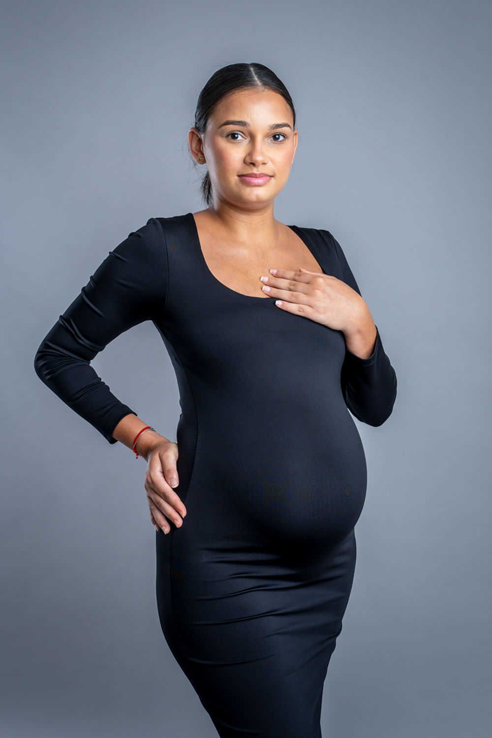 Une femme en robe noire ajustée à manches longues pose sur un fond gris montrant une silhouette enceinte.