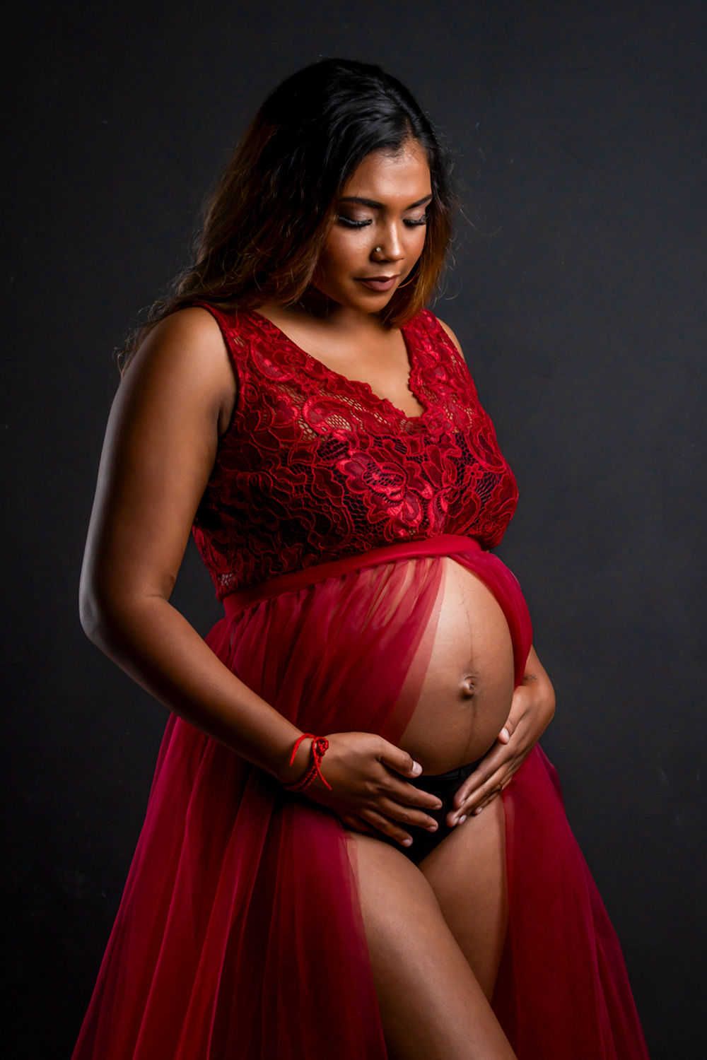 Belle photo de maternité mettant en vedette une future maman dans une robe fluide en dentelle rouge sur un fond sombre.
