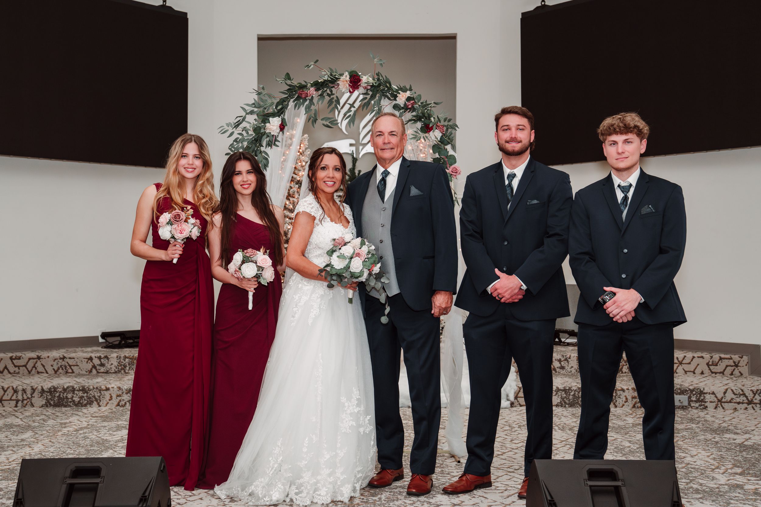 Wedding party in formal attire poses together in front of a floral arch at an indoor venue.