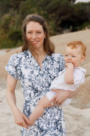 A mother in a blue and white floral dress holds a small baby wearing white against an outdoor backdrop.