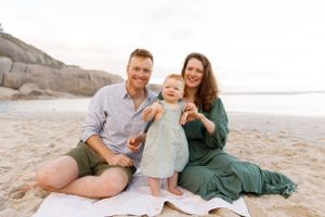 Family enjoys a relaxed moment together on a sandy beach blanket against coastal rocks and ocean.