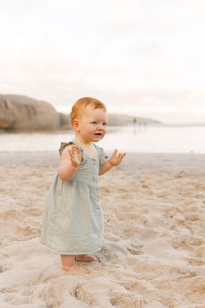 A small toddler wearing a light blue dress stands barefoot on a sandy beach at sunset.