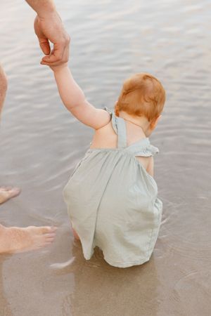 A small child in a light gray romper holds hands with an adult while playing at the water's edge.