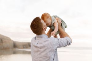 A loving moment between parent and baby lifted in the air against a soft sunset background at a beach.