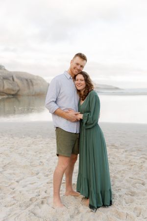 A couple embraces on a sandy beach with rocky cliffs and ocean in the background.