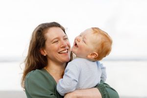A parent and young child share a joyful laughing moment together outdoors against a bright background.