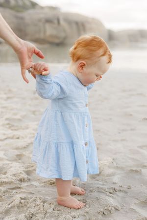 A toddler in a light blue dress takes first steps on the beach while holding a hand.