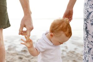 A young child in white clothing holds hands with adults while walking on a sandy beach in bright sunlight.