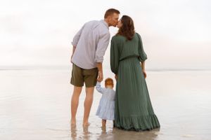 A couple and small child standing together at the beach during sunset while wearing casual summer clothing.