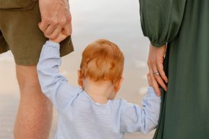 A close-up view of a toddler in blue holding hands with adults on either side while standing on sand.