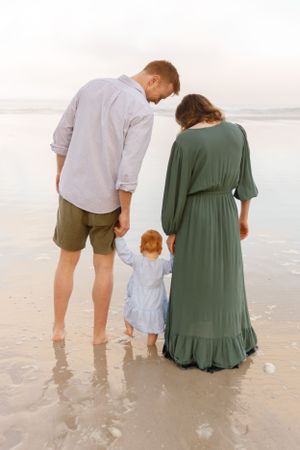 A family of three stands together on a beach at sunset, with a young child holding their hands in the middle.