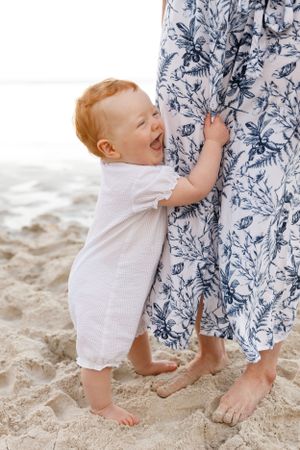 Happy baby in white romper holds onto blue floral dress while standing barefoot on beach sand together.