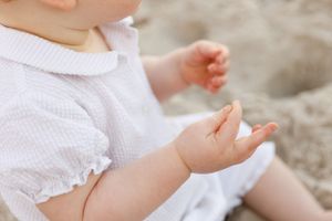 Small hands in white lace dress touch sand while playing outside on a sunny day.