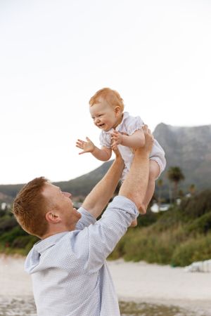 A parent lifts their baby while playing together on a beach with mountains in the background.