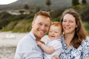 A happy family of three smiles together while sitting near water with mountains in the background.