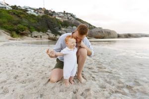 Two people share a tender moment on a sandy beach with rocky cliffs and coastal homes in the background.