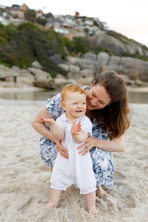 A tender beach moment between parent and toddler in white romper against coastal rock formations at sunset.