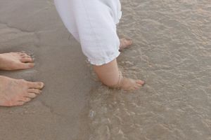 Bare feet and white pants standing in shallow ocean water on a sandy beach.