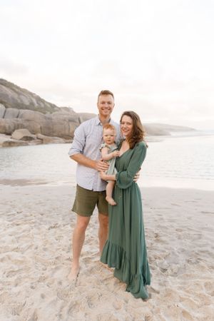 A family poses together on a scenic beach with rocky cliffs in the background at sunset.