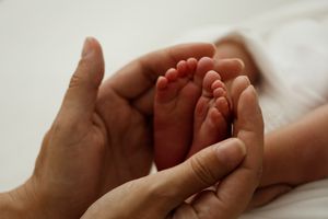 Hands gently cradle a newborn baby's tiny feet and fingers in a tender, intimate close-up photograph.