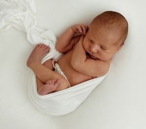 Newborn baby sleeping peacefully wrapped in soft white fabric against a light background.