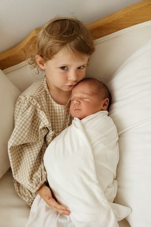 A tender moment shows a young child in a gingham dress holding a swaddled newborn baby on white bedding.