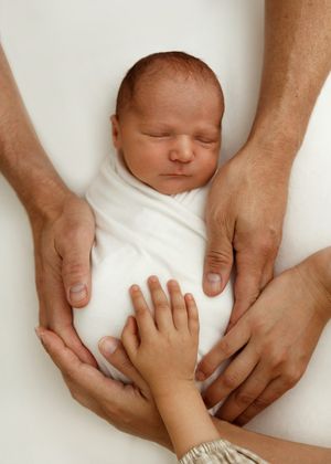 A sleeping newborn baby swaddled in white with multiple hands gently cradling them from above.