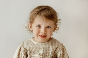 A toddler wearing a gingham dress and cardigan smiles while standing against a light background.