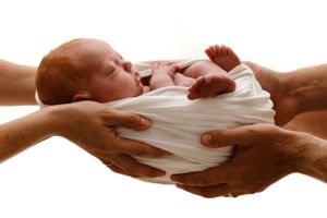 Four adult hands gently cradle and support a swaddled newborn baby in white cloth against white background.