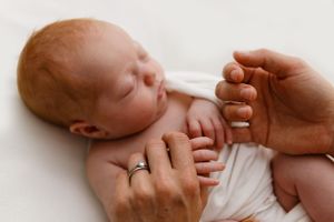 Adult hands gently hold a sleeping newborn baby wearing a white diaper against a light background.