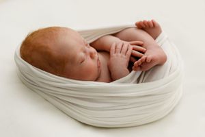 Peaceful newborn baby sleeping while wrapped in soft white fabric against light background.