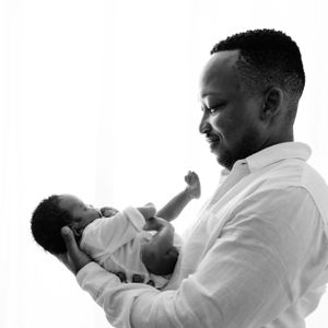 Black and white photo of a tender moment between a father in white shirt and baby in candid pose against light background.