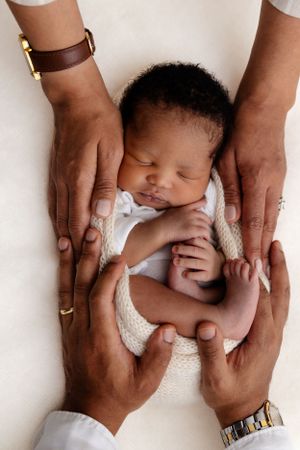 Adult hands wearing jewelry tenderly cradling a sleeping newborn wrapped in white fabric.