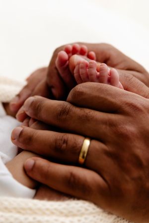 Close-up of adult hands wearing a gold wedding band gently holding tiny baby feet against white fabric.