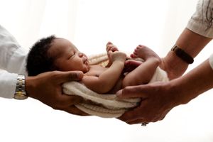 Adult hands wearing watches delicately holding a newborn baby swaddled in beige fabric.
