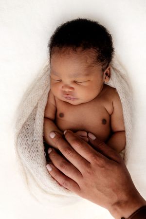 Gentle hands cradle a peaceful newborn baby wrapped in soft white knit fabric.