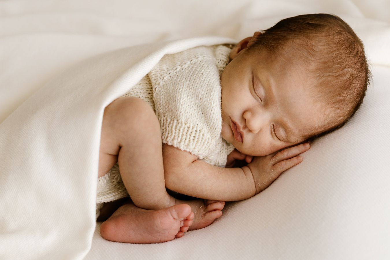 Peaceful newborn baby sleeping curled up under a soft white blanket in a cozy newborn photography pose.