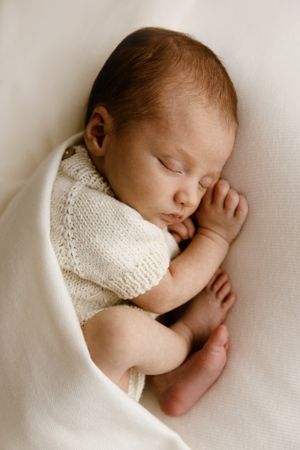 Peaceful newborn baby sleeping curled up in a soft white knit outfit and blanket.
