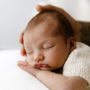A peaceful newborn baby sleeping on white fabric, cradled gently by an adult's tattooed hand.
