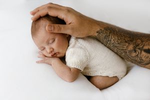 Sleeping newborn baby in cream knit outfit cradled by tattooed father's hand on white background.