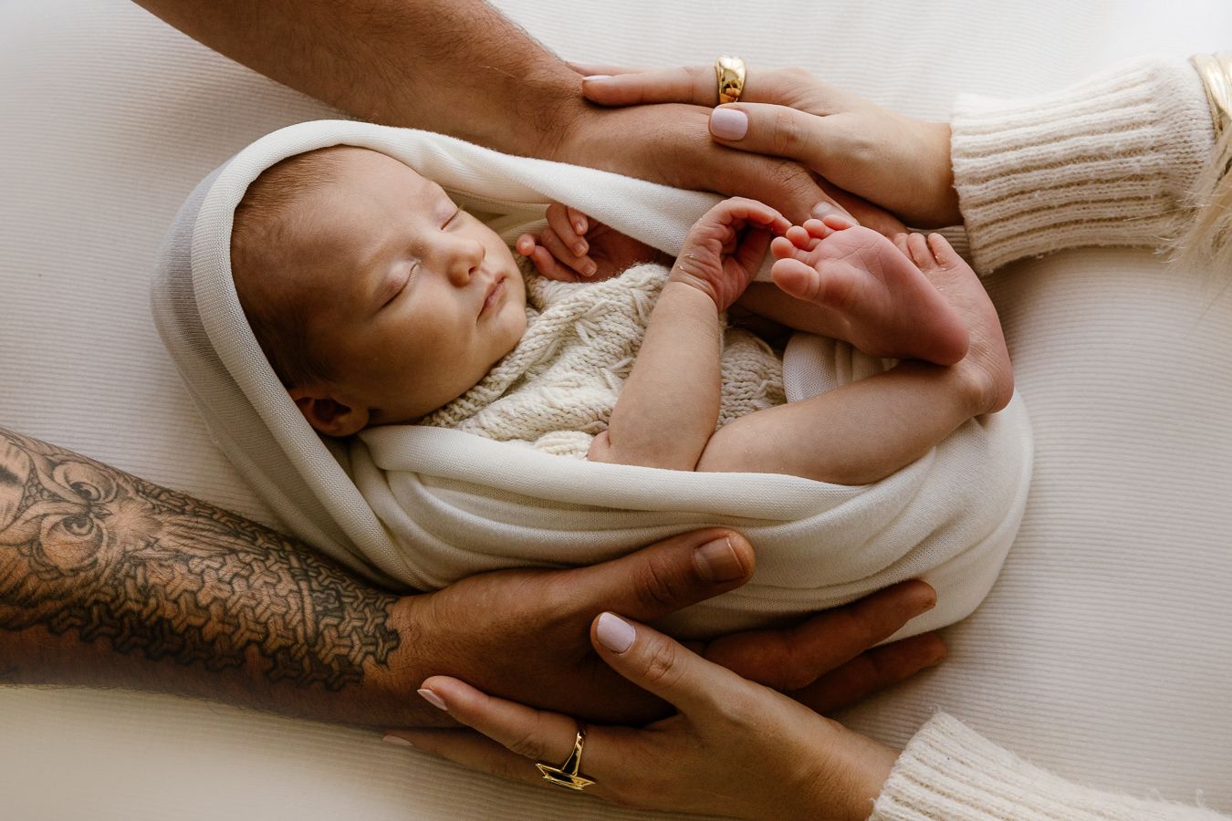 Parents cradling their sleeping newborn baby wrapped in a white knit blanket, showing tattooed and ringed hands.