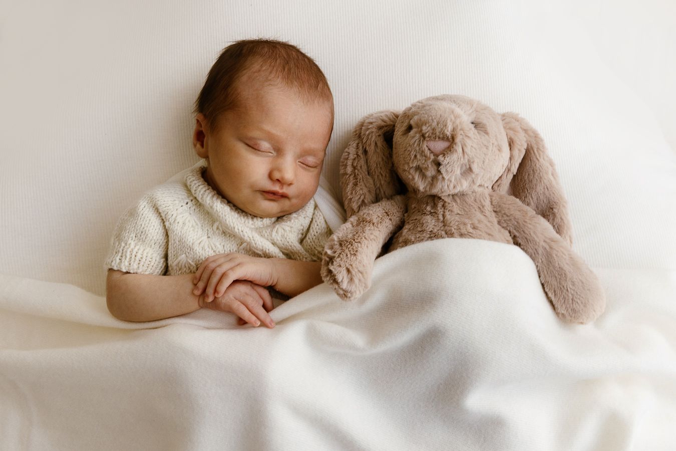 Peaceful newborn baby sleeping beside a soft plush bunny rabbit toy, wrapped in a cozy white knit blanket.