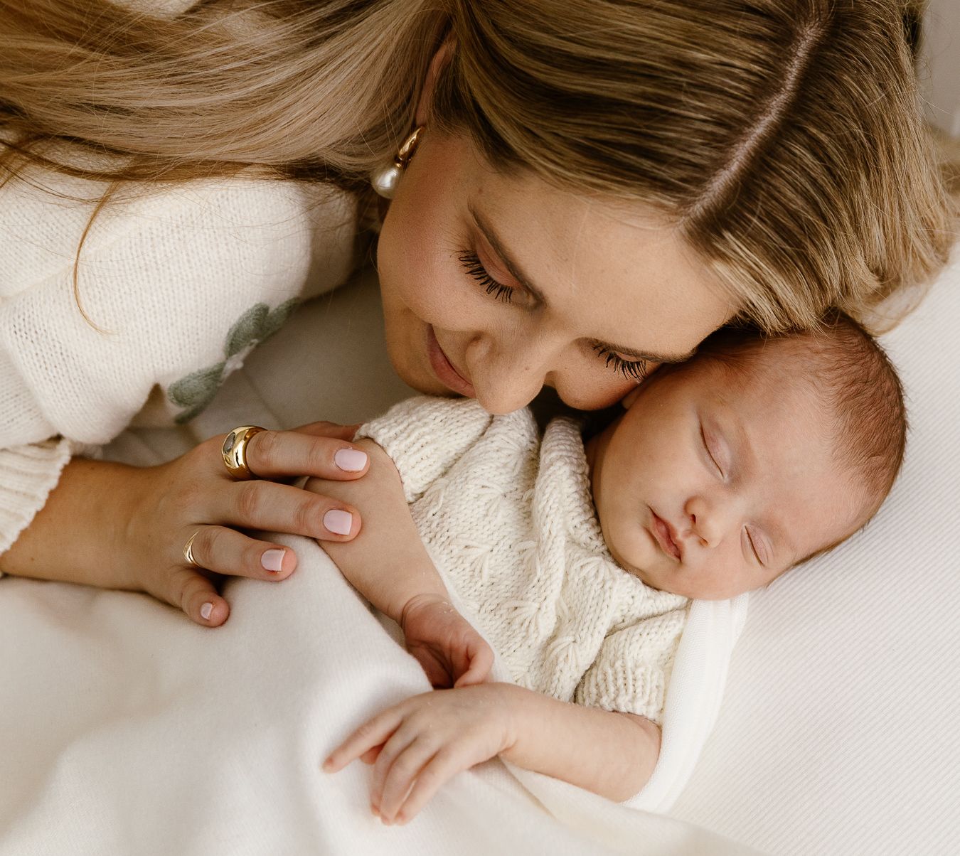 A tender mother gently kisses her sleeping newborn baby wrapped in a cream knit blanket.