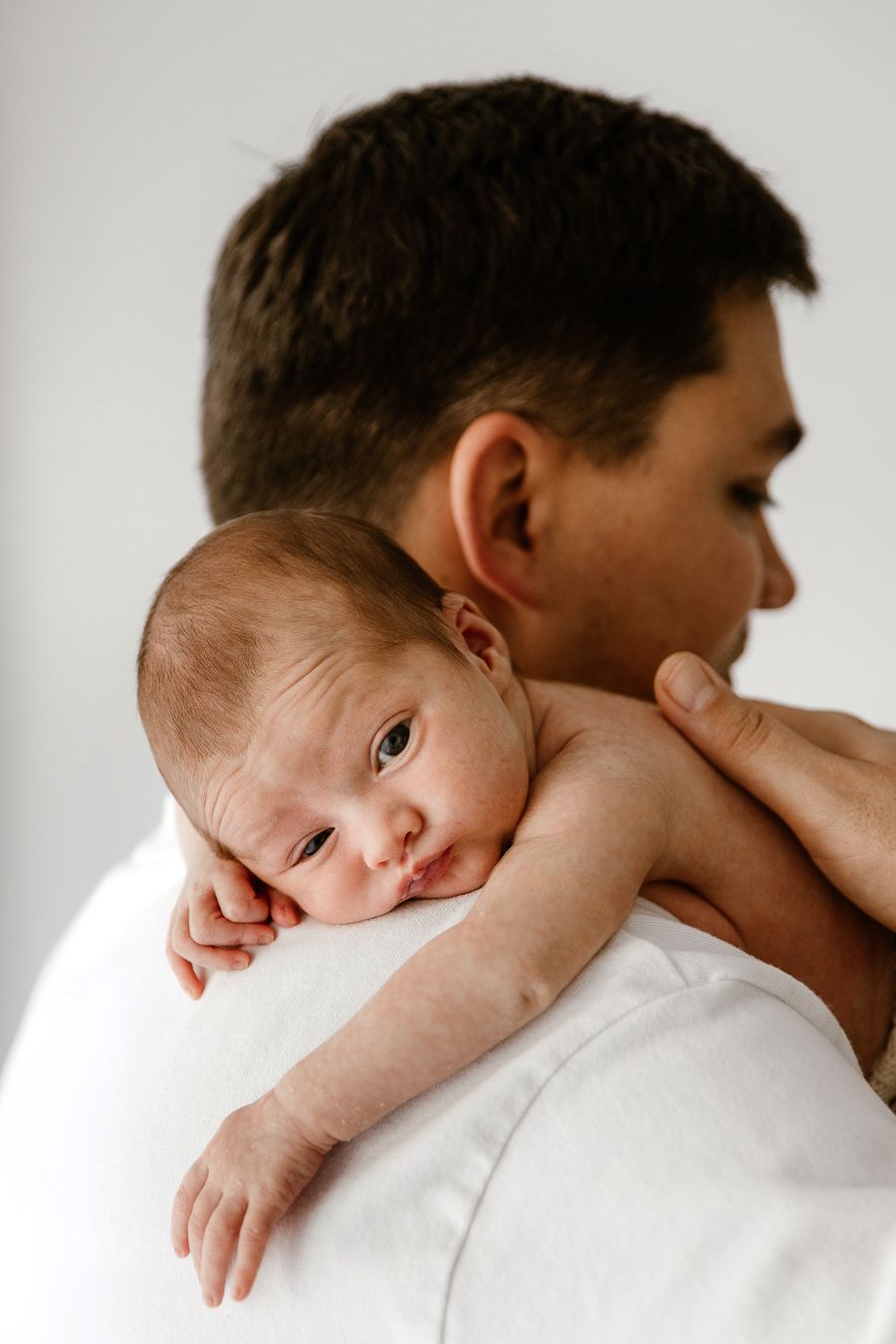 A father in a white shirt gently holds his newborn baby on his shoulder against a soft white background.